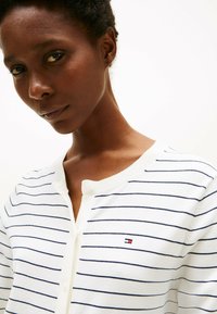 Cardigan bianco a righe con linee orizzontali blu navy, dotato di scollatura rotonda e chiusura con bottoni. Piccolo logo sulla parte sinistra del petto.