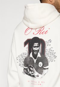 Sudadera blanca con un gráfico en blanco y negro de un hombre sosteniendo un zapato, con acentos rojos y texto. Detalles notables incluyen "O Rei" y "10".