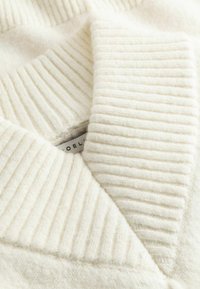 Gros plan sur un tissu tricoté doux, côtelé, de couleur crème avec une étiquette visible montrant partiellement le nom de la marque.