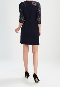 Robe bleu marine avec manches en dentelle trois-quarts, corsage ajusté et longueur courte. Des chaussures à talons noires complètent la tenue.