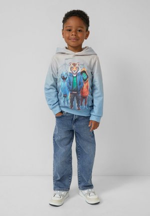 Jongentje in een lichtblauwe hoodie met dierenfiguren en een blauwe spijkerbroek, staand tegen een effen achtergrond.