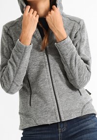 Osoba nosząca szary polar z zamkiem, trzymająca ręce w pobliżu kaptura, w połączeniu z ciemnoniebieskimi dżinsami na białym tle.