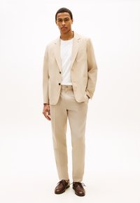 Νέος άνδρας ντυμένος με μπεζ κοστούμι, λευκό t-shirt και καφέ παπούτσια, στέκεται μπροστά σε απλό λευκό φόντο.