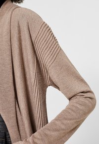 Beżowy sweter kardigan z dzianiny z prążkowanymi detalami na rękawach, miękką fakturą oraz otwartym przodem o luźnym kroju.