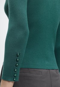 Πράσινο ριμπ τοπ με μακριά μανίκια και έξι διακοσμητικά κουμπιά στη μανσέτα, σε συνδυασμό με σκούρο γκρι παντελόνι με ραφές.