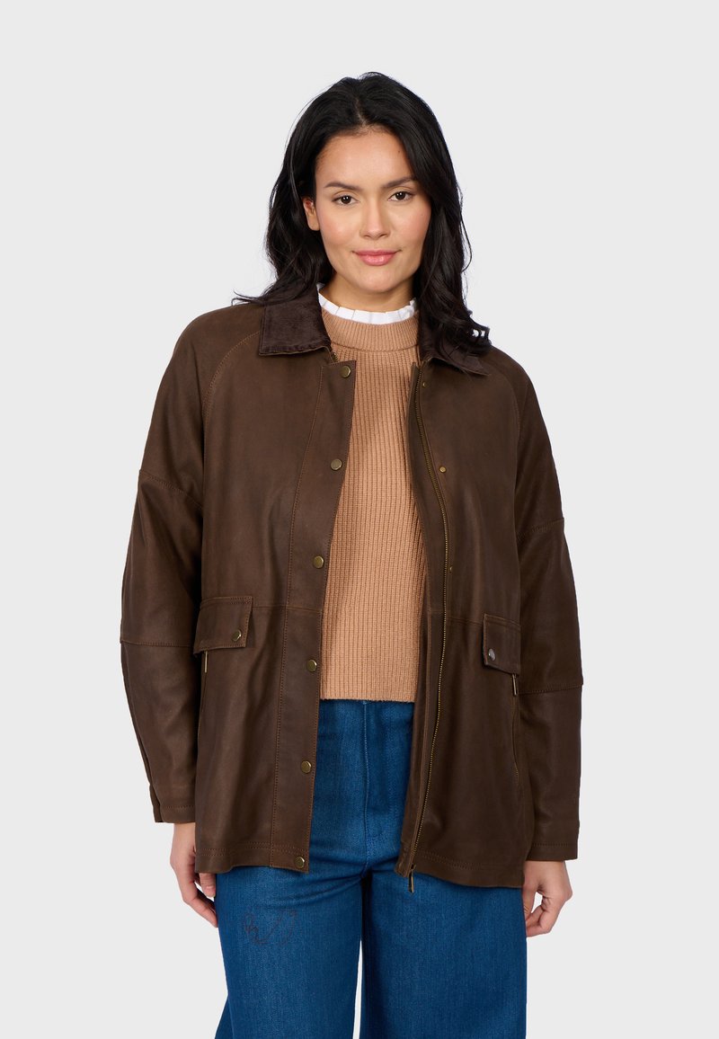 Kobieta z ciemnymi włosami nosząca brązową skórzaną kurtkę na beżowym swetrze i niebieskich dżinsach, stojąca na jednolitym jasnym tle.