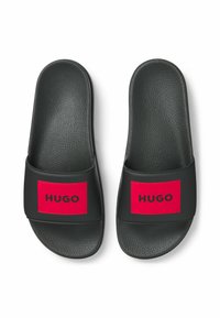 Musta liukusandaalit tekstuuripinnalla, leveä hihna, jossa on punainen suorakulmainen lappu, jossa lukee "HUGO" paksuilla mustilla kirjaimilla.