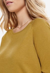 Senapsgul stickad tröja med bred rund halsringning och raglanärmar. Mjuk textur och detaljerad sömnad vid axelsömmarna.