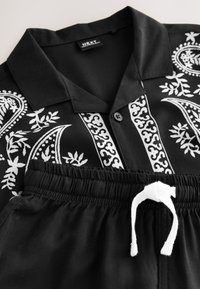 Camisa negra de manga corta con patrones florales y abstractos blancos combinada con pantalones negros de cintura elástica que cuentan con un cordón blanco.