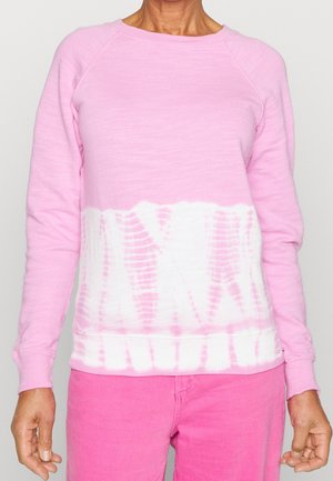 Osoba ubrana w różowo-biały sweatshirt z długim rękawem w stylu tie-dye oraz jasnoróżowe spodnie, stojąca na tle jednolitego, jasnego tła.