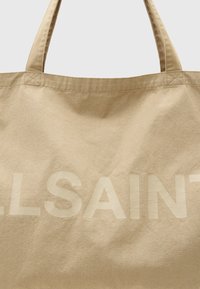 Beige Canvas-Umhängetasche mit großem eingeprägtem "ALLSAINTS"-Text. Verfügt über robuste, lange Tragegriffe und eine weiche Textur. Einfaches, minimalistisches Design.