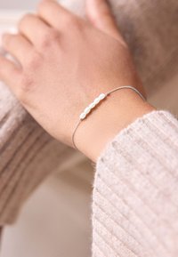 Silver bracelet featuring five round, white beads, worn on a hand with a soft beige sweater in the background.