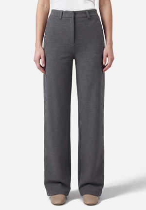 Donna che indossa pantaloni grigi a vita alta e gamba larga abbinati a scarpe beige, in piedi davanti a uno sfondo bianco uniforme.