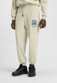 Crème-kleurige joggingsbroek van zacht materiaal met een elastische tailleband, voorzien van een blauw logo en afbeelding op de linker dij. Gecombineerd met zwarte sneakers.