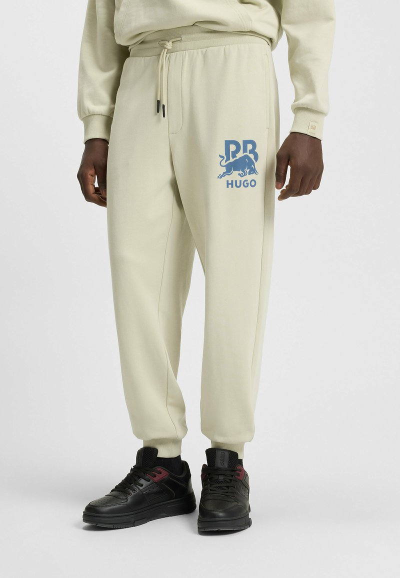Crème-kleurige joggingsbroek van zacht materiaal met een elastische tailleband, voorzien van een blauw logo en afbeelding op de linker dij. Gecombineerd met zwarte sneakers.