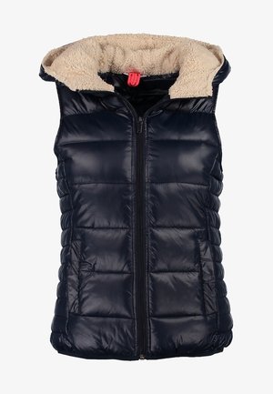 Donna che indossa jeans aderenti blu, stivaletti neri lucidi alla caviglia, maglia grigia a maniche lunghe, gilet imbottito blu scuro con fodera in pile, porta uno zaino nero.