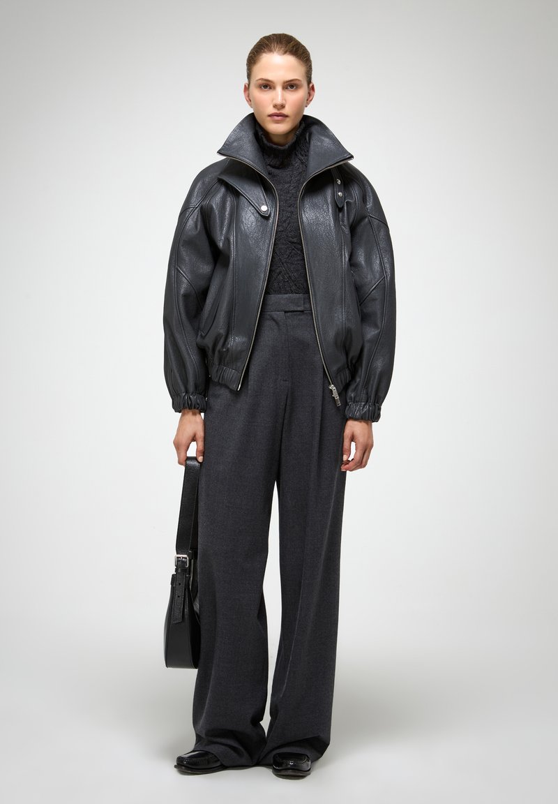 Schwarze Lederbomberjacke mit hohem Kragen, kombiniert mit weit geschnittenen grauen Hose. Das Model hält eine schwarze Umhängetasche und steht vor einem neutralen Hintergrund.