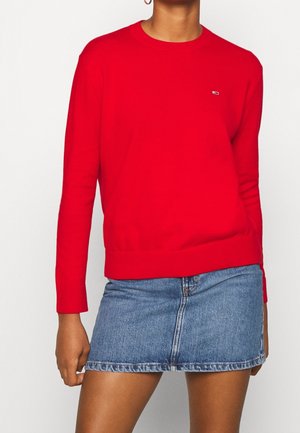 Person iført en knaldrød langærmet sweater med lille logo på brystet og en blå denim mininederdel mod en ensfarvet baggrund.
