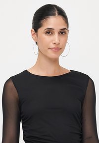 Črna majica z dolgimi, prosojnimi rokavi, okroglo izrezom in prilegajočo silhueto. Model nosi velike uhane iz krogov. Gladka tekstura, minimalističen dizajn.