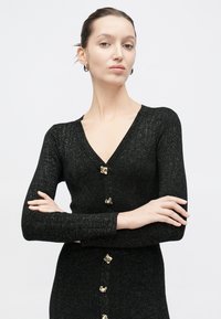 Γυναίκα με μαλλιά δεμένα πίσω, που φοράει μαύρη ζακέτα με ριμπ ύφανση και χρυσά κουμπιά, καθώς και ασημένια σκουλαρίκια κρίκους, με σταυρωμένα χέρια και λευκό φόντο.