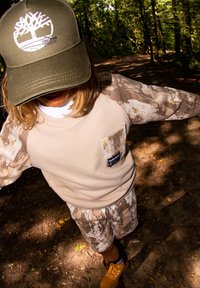 Sweatshirt beige avec des manches en camouflage, détail de poche et logo. Shorts camouflage assortis. Casquette olive avec logo. Bottes jaunes.