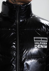 Černá péřová bunda s lesklým povrchem, vysokým límcem, plným zipem a bílým logem "HILFIGER DENIM" na levé hrudi.