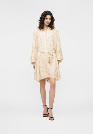 Kobieta o kręconych włosach, ubrana w długą sukienkę z długim rękawem w jasnobeżowym kolorze z złotymi wzorami oraz czarne sandały na wysokim obcasie, stojąca na białym tle.