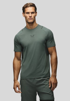 Homme musclé debout face à l'avant, portant un t-shirt de sport ajusté manches courtes vert foncé et un short assorti sur un fond gris.