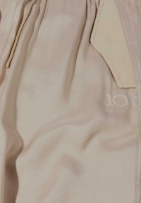 Beige stof met een gladde textuur, voorzien van een geborduurd "10" en een zakdetail. De stof heeft een subtiele glans en valt mooi.