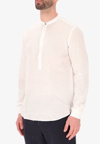 Camicia bianca in lino a maniche lunghe con colletto alla coreana, patta con bottoni e orlo arrotondato. Il tessuto sembra leggero e texturizzato.