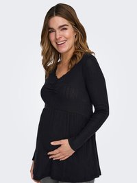 Haut de maternité noir à manches longues avec texture côtelée, fronces au niveau de la poitrine et ourlet évasé ; le modèle tient doucement son ventre.