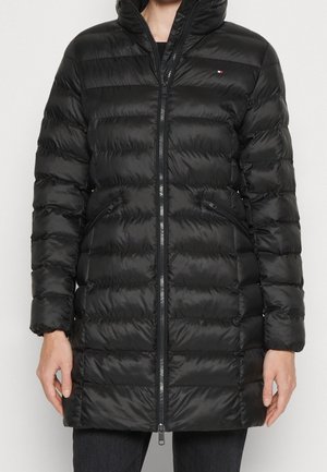 Femme portant un manteau matelassé noir avec un col monté, une fermeture éclair à l'avant, des poches zippées et un petit logo sur la poitrine.