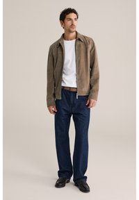 Brūngans zamšādas žakete ar rāvējslēdzēju priekšpusē, balts t-krekls un tumši zili plašām kājām džinsi. Melni apavi papildina apģērbu. Vienkāršs fons.