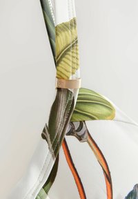 Weißer Stoff-BH-Träger mit grünen Pflanzenmustern, goldenen Hardware-Akzenten und sichtbaren Nähdetails.