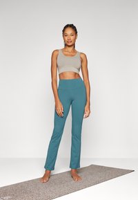 Világosszürke anyagú melltartó felső, fitted türkiz leggings-szel párosítva, mindkettő sima textúrájú. A modell egy textúrált szőnyegen áll.