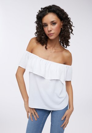 Top bianco off-the-shoulder con design a volant, realizzato in tessuto leggero; abbinato a jeans blu. Texture morbida e vestibilità casual.