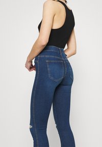 Vaqueros skinny de tiro alto en denim azul oscuro con bolsillos traseros. Presentan rodillas desgastadas y costuras en contraste a lo largo de las costuras.