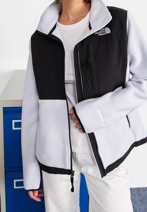 Persona que lleva una chaqueta polar con cremallera en blanco y negro de The North Face sobre una camisa blanca y pantalones blancos, de pie cerca de archivadores azules.