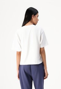 Weißes kurzärmliges T-Shirt mit lockerer Passform und Rundhalsausschnitt, kombiniert mit dunkelblauen Hosen, von hinten betrachtet. Glatte Stoffstruktur.