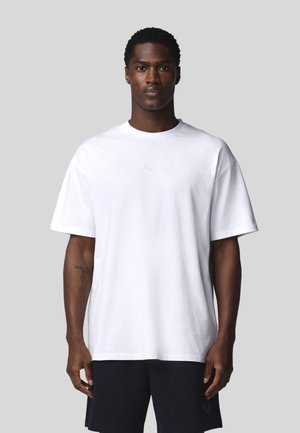 Homme portant un t-shirt blanc oversize uni et un short noir, debout devant un fond gris uni.