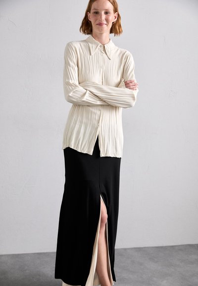 Cream, pleated long-sleeve shirt with button closure paired with a fitted black skirt featuring a thigh-high slit. Smooth texture, modern silhouette.