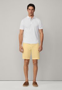 White polo shirt, yellow shorts, brown shoes. Polo features a classic collar and soft fabric. Shorts have a simple design and relaxed fit.