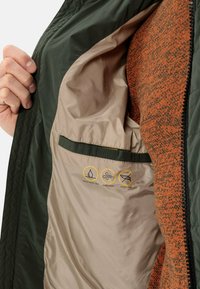 Indersiden af en mørkegrøn jakke med en beige foring; har ikoner, der viser vandafvisende, vindtæt og dunfri egenskaber.