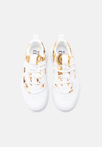 Λευκά αθλητικά παπούτσια με υφή και χρυσό σχεδιασμό στο επάνω μέρος και χοντρές λαστιχένιες σόλες· κορδόνια και λουριά στο ίδιο λευκό χρώμα.