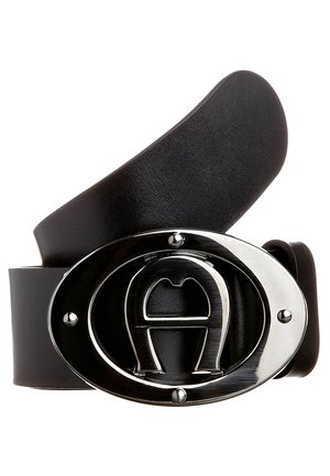 Ceinture en cuir noir avec une boucle argentée ovale brillante présentant un "A" stylisé en relief au centre, enroulée sur un fond blanc.