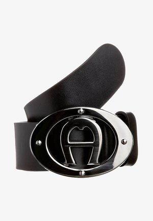 Ceinture en cuir noir avec une boucle argentée ovale brillante présentant un "A" stylisé en relief au centre, enroulée sur un fond blanc.