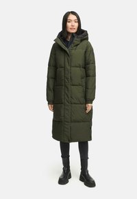 Lang grøn puffjakke med hætte, featuring et kasseformet design, knaplukning foran og elastiske manchetter. Båret over sorte jeans og kraftige sorte støvler.