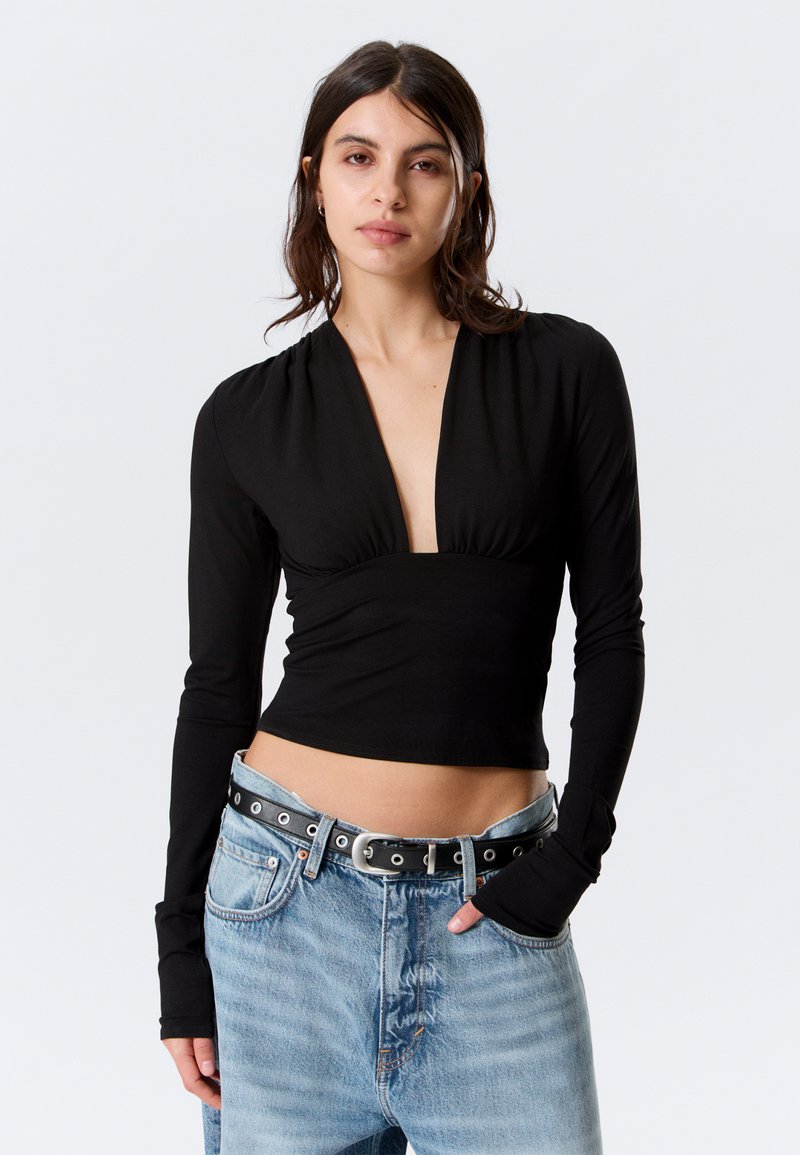 Femme aux cheveux foncés mi-longs portant un haut noir à manches longues avec un décolleté plongeant en V et un jean bleu clair avec une ceinture noire.