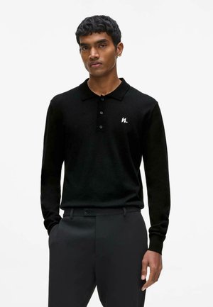 Mees kannab musta pika varrukaga polo särke, millel on kolm nööpi ja valge tikandiga "KL" logo rinnal, käed taskutes.