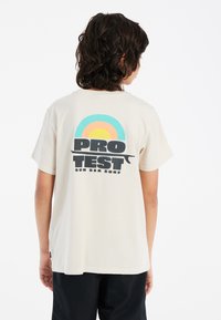 Šviesiai rudos spalvos medvilnės marškinėliai su grafiniu dizainu, kuriame mėlyni, oranžiniai ir geltoni pusmėnuliai virš žodžio "PRO TEST" parašyto drąsiais juodais rašmenimis.